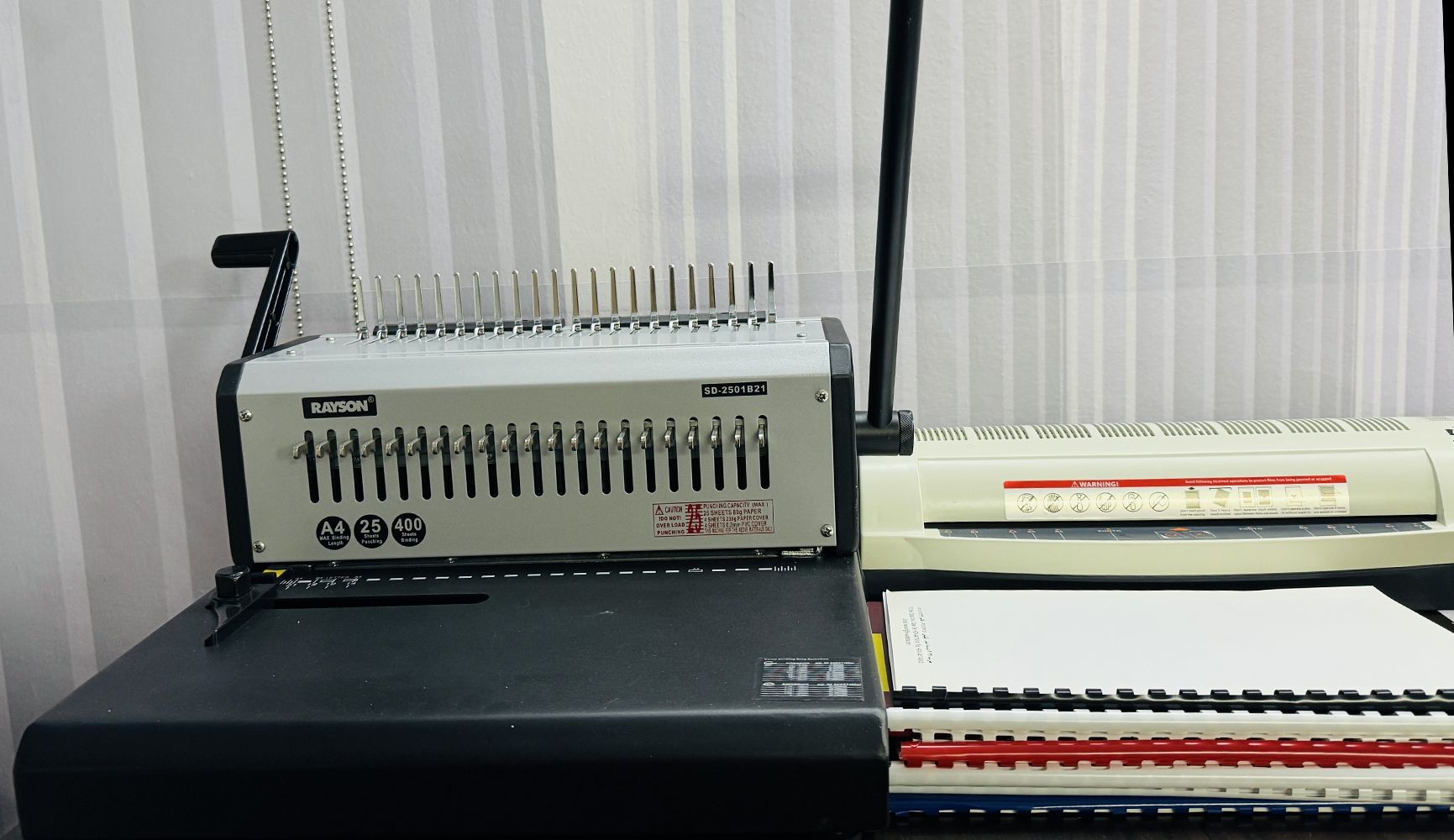 Binding machine on a desk with combs and lever, next to a laminated sheet stack.